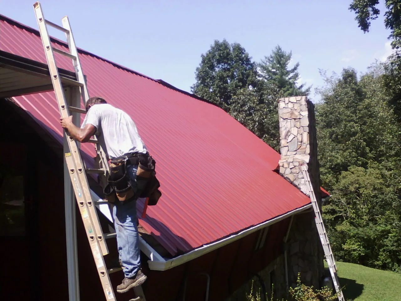 Roofer on a ladder beside a red metal roof and stone chimney — Soffit & Fascia Repair by Roofing Brothers in Courtland